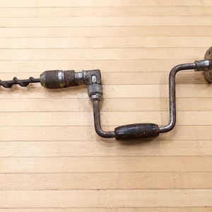 May include: A vintage hand brace drill featuring a dark metal frame with a rusted finish, a spiral auger bit attached to the chuck, and two dark brown wooden handles located on the central crank and the end knob for manual operation.