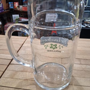 May include: A clear glass beer mug featuring a sturdy handle and a thick base. The front of the glass displays the Heineken logo in white and green, with a small red star above the text and the word Holland printed in black below the logo.