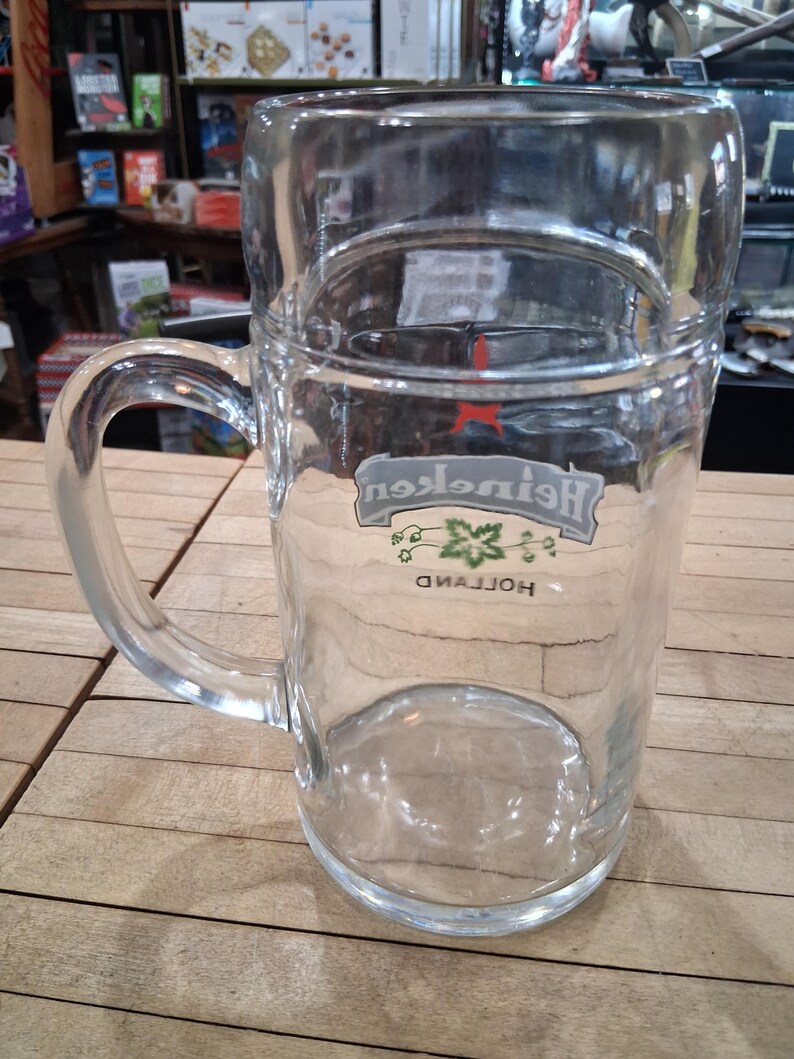 May include: A clear glass beer mug featuring a sturdy handle and a thick base. The front of the glass displays the Heineken logo in white and green, with a small red star above the text and the word Holland printed in black below the logo.