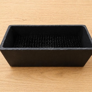 May include: A rectangular black flower frog, also known as a kenzan, with a dense array of black pins. The frog is made of a dark material and is designed to hold flower stems in place for floral arrangements.