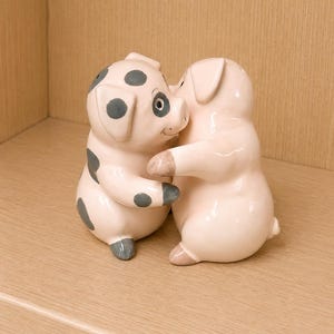 May include: Two ceramic pig figurines in a hugging pose. The left pig is light pink with dark gray spots on its head, ears, and body. The right pig is solid light pink with dark gray accents on its hooves. Both figures have smooth, glossy finishes.