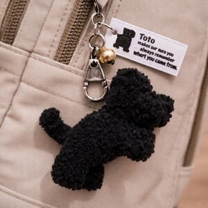 May include: A black plush dog keychain with a silver clasp and a gold bell charm. The keychain is attached to a beige bag. A small white tag reads "Toto makes sure you always remember whert you came from."