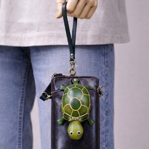 May include: A black leather crossbody bag with a green turtle design. The turtle has a detailed shell pattern and is attached to the front of the bag. The bag has a dark green strap and a gold-colored chain.