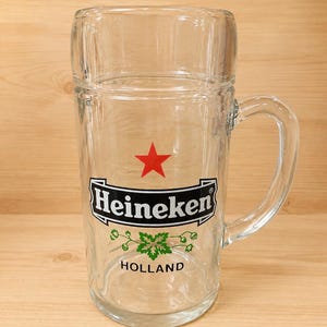 May include: A clear glass beer mug featuring the Heineken logo with a red star, the brand name in black text, a green hop graphic, and the word HOLLAND printed below. The mug has a sturdy handle and a smooth, cylindrical body design.