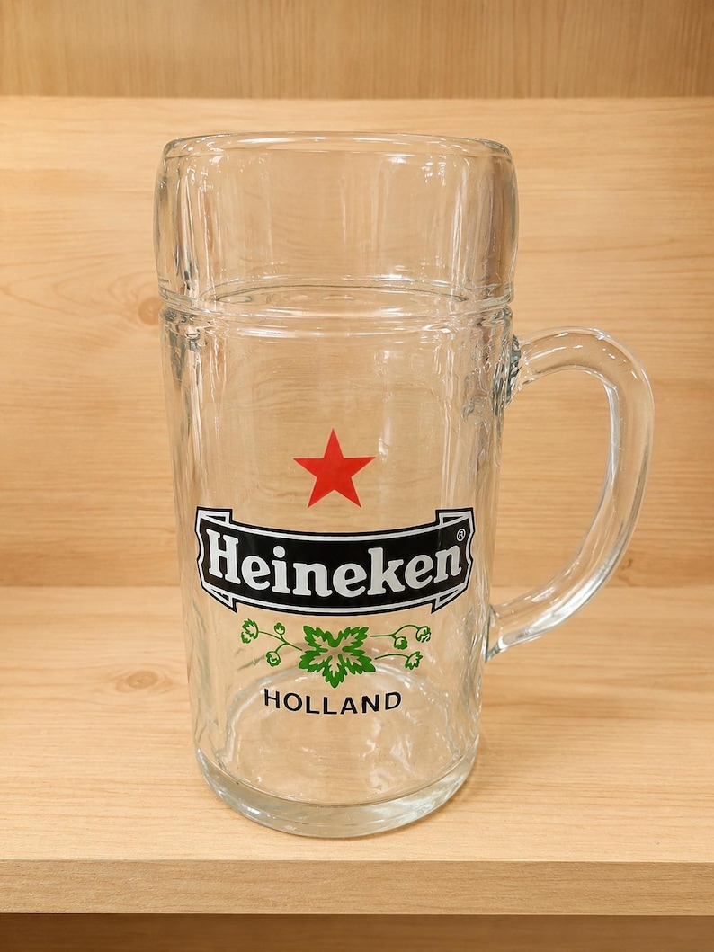 May include: A clear glass beer mug featuring the Heineken logo with a red star, the brand name in black text, a green hop graphic, and the word HOLLAND printed below. The mug has a sturdy handle and a smooth, cylindrical body design.