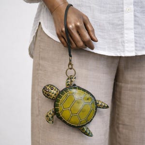 May include: A turtle-shaped coin purse with a yellow and green design. The purse has a black strap and a gold-colored clasp. The turtle's shell features a geometric pattern. The purse is held against a background of tan pants and a white shirt.