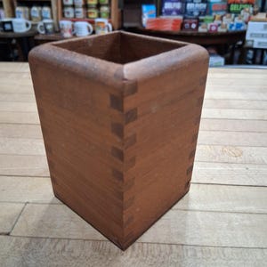 May include: A square wooden box with a warm brown finish. The box is constructed with visible dovetail joints at the corners, showcasing the craftsmanship. The box is empty and sits on a light-colored wooden surface.