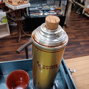 May include: Vintage gold and silver vacuum bottle with a cork stopper. The bottle has the words "Vacuum Bottle" printed on it. A small, red cup sits next to the bottle. The bottle is in a blue tray.