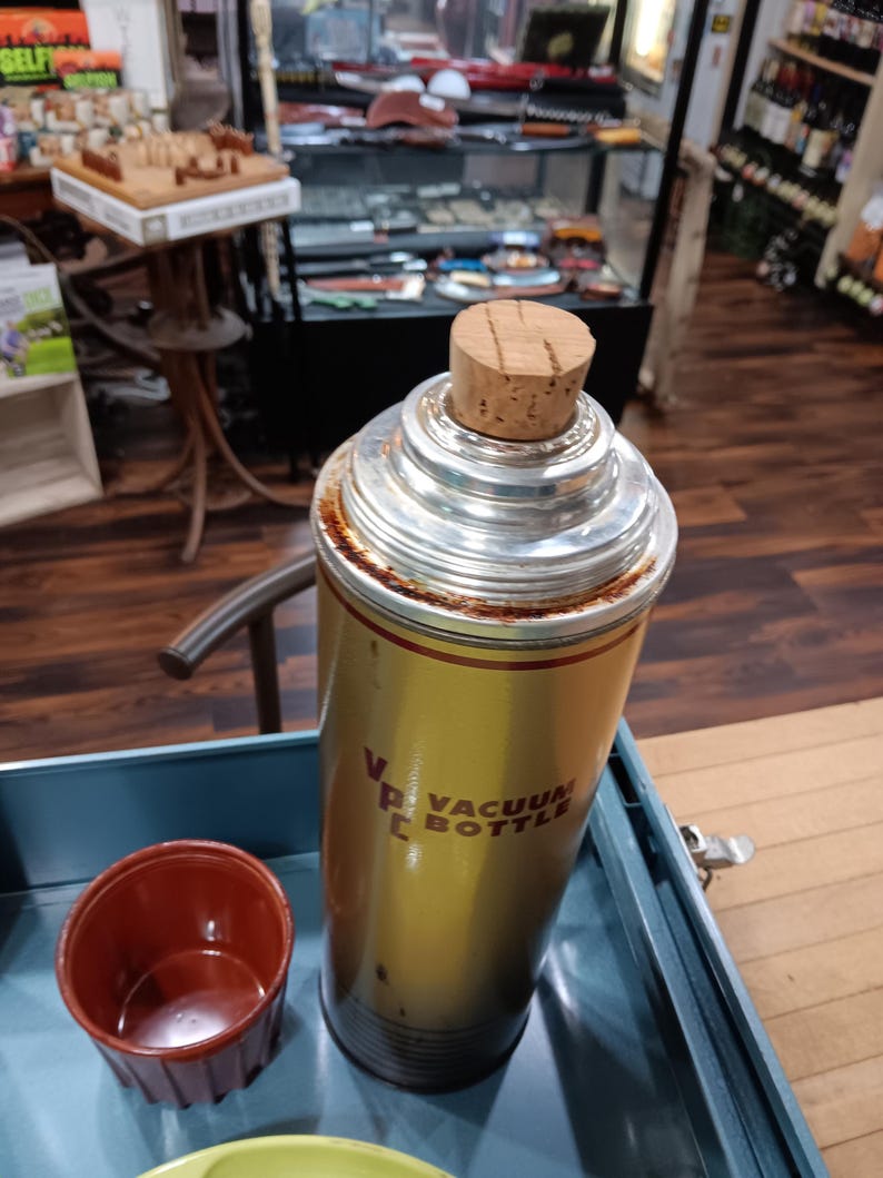 May include: Vintage gold and silver vacuum bottle with a cork stopper. The bottle has the words "Vacuum Bottle" printed on it. A small, red cup sits next to the bottle. The bottle is in a blue tray.