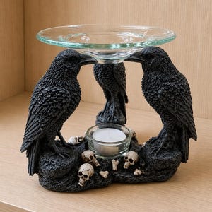 May include: A decorative black raven candle holder with a clear glass dish. The base features skulls and bones, with a small glass cup holding a white candle. The ravens are perched on a textured base.