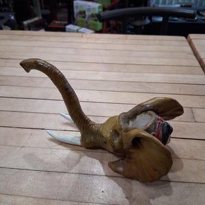 May include: A detailed sculpture of an elephant head, featuring a long, curved trunk and large ears. The sculpture is primarily brown and tan, with white tusks. The elephant head is lying on a wooden surface.