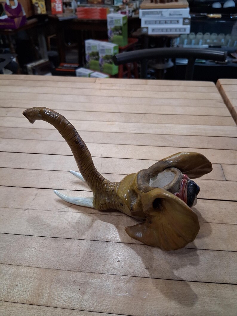 May include: A detailed sculpture of an elephant head, featuring a long, curved trunk and large ears. The sculpture is primarily brown and tan, with white tusks. The elephant head is lying on a wooden surface.