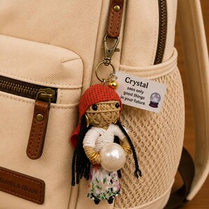 May include: A handmade crochet doll keychain with a red headscarf, white top, and floral skirt, holding a pearl. The doll hangs from a beige backpack with brown leather accents. A tag reads "Crystal sees only good things in your future."