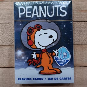 May include: A deck of Peanuts playing cards featuring Snoopy in an orange spacesuit with red goggles. The box has a blue background with stars and the text "PEANUTS" at the top. The bottom of the box reads "PLAYING CARDS • JEU DE CARTES".
