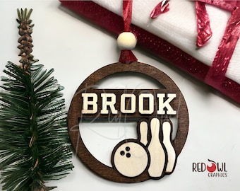 May include: A wooden Christmas ornament with the name "BROOK" in gold letters and a bowling ball and pins design. The ornament is brown with a white background.