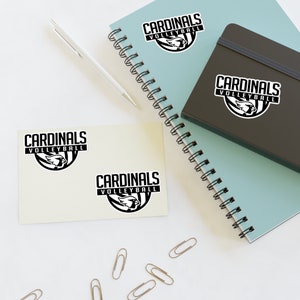 May include: Two beige notepads with black and white stickers that say "Cardinals Volleyball". The stickers feature a cardinal bird graphic. The notepads are on a white surface with a blue spiral notebook and a black notebook.