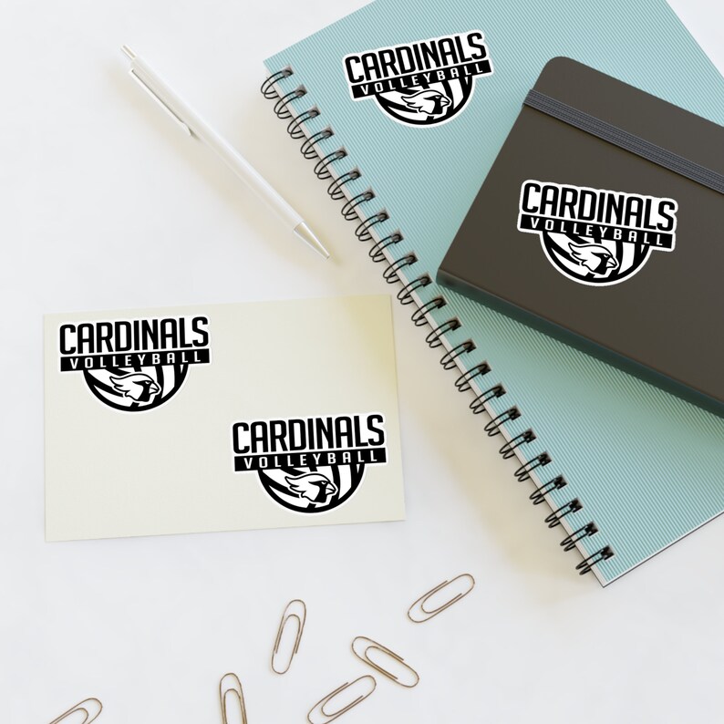 May include: Two beige notepads with black and white stickers that say "Cardinals Volleyball". The stickers feature a cardinal bird graphic. The notepads are on a white surface with a blue spiral notebook and a black notebook.