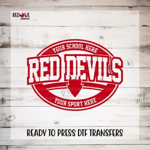 May include: A red and white graphic design featuring the text "RED DEVILS" in bold, block letters, with "YOUR SCHOOL HERE" and "YOUR SPORT HERE" in a circular banner. A devil head graphic is in the center. The words "READY TO PRESS DTF TRANSFERS" are at the bottom.