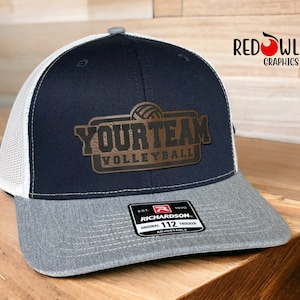 May include: A navy blue and gray trucker hat with a white mesh back. The hat has a brown leather patch with the text "YOUR TEAM VOLLEYBALL" and a volleyball graphic.