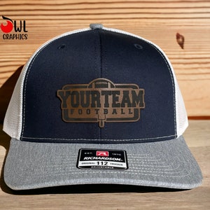 May include: A navy blue and white trucker hat with a brown leather patch that says "YOUR TEAM FOOTBALL" with a football graphic. The hat is adjustable and has a gray underside.