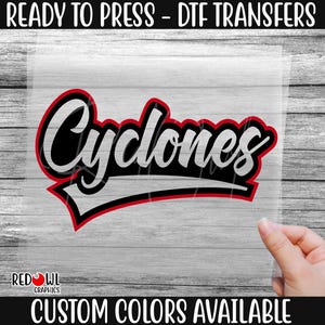 May include: A white iron-on transfer sheet with the word "Cyclones" in black lettering with a red outline. The text is in a bold, stylized font. The transfer sheet is ready to be pressed onto fabric.