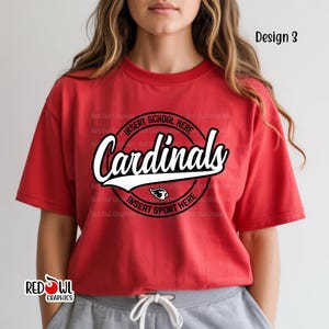 May include: Red t-shirt with the word "Cardinals" in a white script font, outlined in black, above a white baseball bat graphic. The shirt has a circular design with the text "Insert School Here" and "Insert Sport Here".