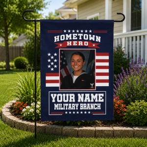 Puede incluir: Bandera de jardín azul marino con el texto "Hometown Hero" y espacio para personalización. Presenta una foto de una persona con uniforme militar, con imágenes de la bandera estadounidense. La bandera se muestra en un soporte de metal negro.