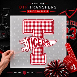May include: A custom DTF transfer sheet featuring a large letter T with a red and white gingham pattern, overlaid with a white pennant graphic containing the word TIGERS in red text. The sheet is held against a black background with sports gear.