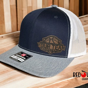 May include: A navy blue and white trucker hat with a brown leather patch featuring a basketball and the text "Your Team Basketball".