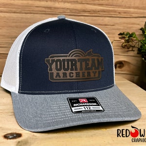 May include: A navy blue and white trucker hat with a brown leather patch that says "Your Team Archery". The hat is adjustable and has a gray mesh back.