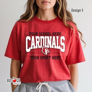 May include: Red t-shirt with the text "CARDINALS" in white block letters. The shirt also features a red cardinal bird logo and the text "YOUR SCHOOL HERE" and "YOUR SPORT HERE". The shirt is a design from Red Owl Graphics.