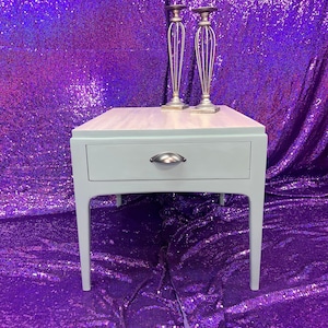 Pu&ograve; includere: Un tavolino da salotto blu chiaro con un cassetto e maniglie in metallo argentato. Il tavolino ha un piano in legno e poggia su quattro gambe. Due portacandele in metallo argentato sono posizionati sul tavolino.