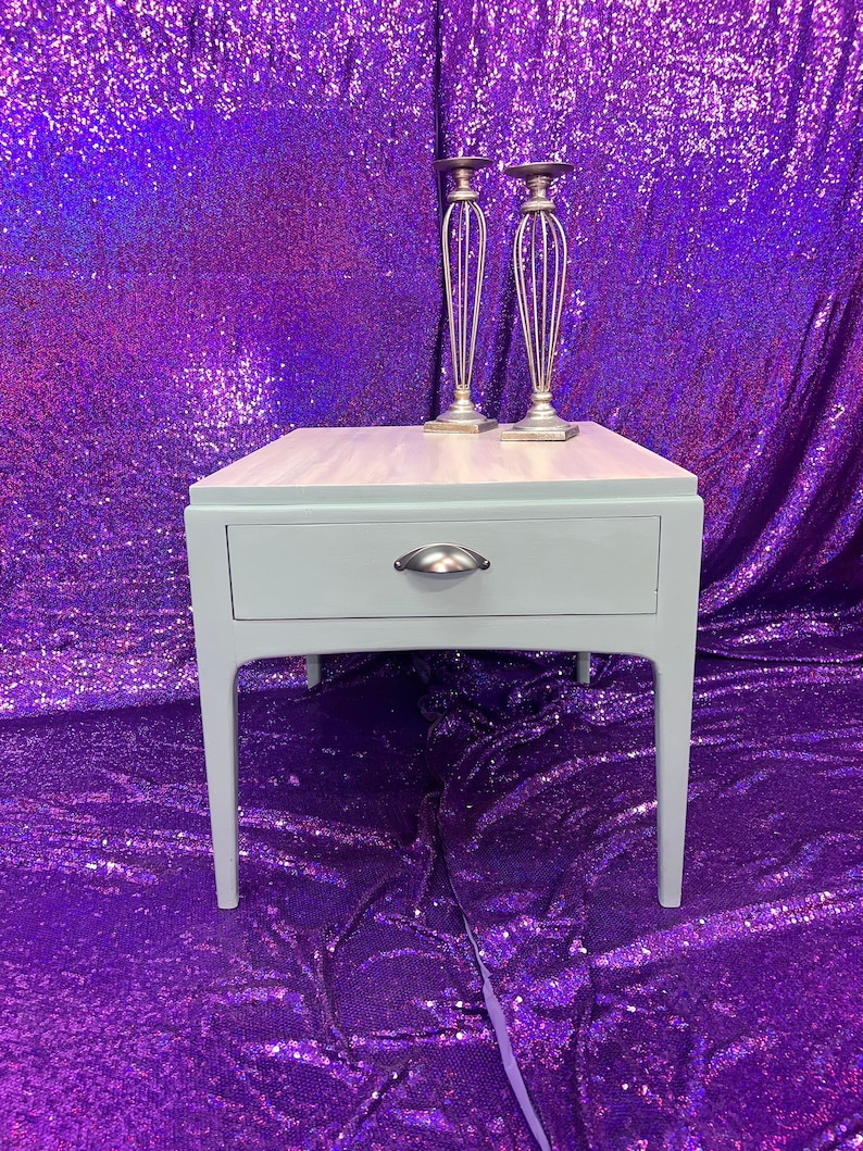 Pu&ograve; includere: Un tavolino da salotto blu chiaro con un cassetto e maniglie in metallo argentato. Il tavolino ha un piano in legno e poggia su quattro gambe. Due portacandele in metallo argentato sono posizionati sul tavolino.