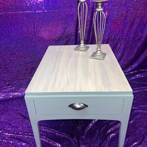Pu&ograve; includere: Un tavolino da salotto blu chiaro con un cassetto e una maniglia argentata. Il tavolo ha un piano in legno sbiancato e si trova su uno sfondo di paillettes viola. Due candelabri argentati sono sul tavolo.