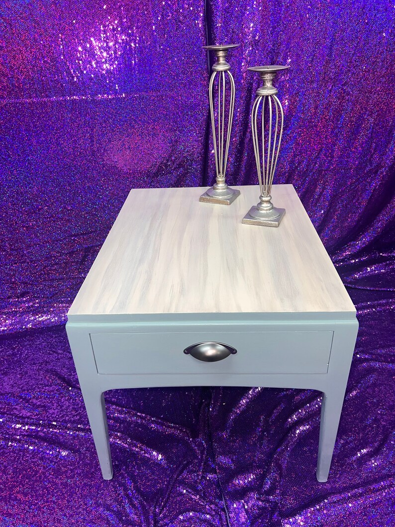 Pu&ograve; includere: Un tavolino da salotto blu chiaro con un cassetto e una maniglia argentata. Il tavolo ha un piano in legno sbiancato e si trova su uno sfondo di paillettes viola. Due candelabri argentati sono sul tavolo.