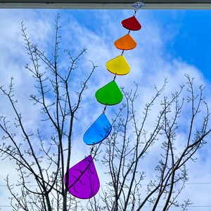 Könnte beinhalten: Ein regenbogenfarbener, tropfenförmiger Windspiel hängt an einem Fenster vor einem blauen Himmel mit weißen Wolken. Das Windspiel ist aus Glas und hat einen violetten, blauen, grünen, gelben, orangen und roten Tropfen.