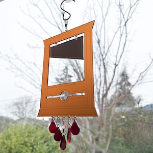 May include: A decorative orange and brown guillotine-shaped wind chime. The wind chime has a rectangular frame with a dark brown blade and a series of red teardrop-shaped pendants hanging from the bottom. It is suspended by a metal hook.