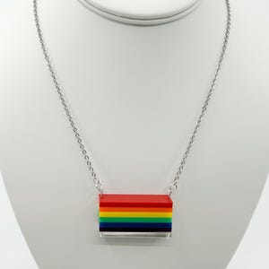 Könnte beinhalten: Halskette mit silberner Kette und rechteckigem Anhänger. Der Anhänger ist ein Stapel bunter Blöcke in den Farben des Regenbogens. Die Halskette wird auf einer weißen Schaufensterpuppe präsentiert.