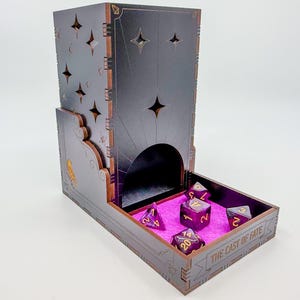 May include: A dark gray wooden dice tower with star-shaped cutouts and a tray. The tray contains several purple dice with gold numbering, resting on a pink felt surface. The text "THE CAST OF FATE" is visible on the tray.