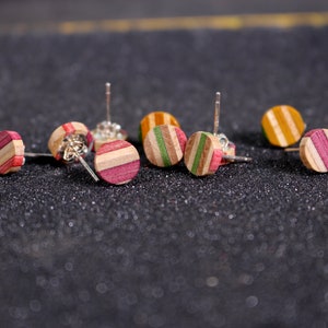 Peut inclure: Plusieurs boucles d'oreilles rondes à clous avec un motif rayé. Les boucles d'oreilles sont en bois et présentent diverses combinaisons de couleurs, notamment des rayures roses, vertes et jaunes. Les boucles d'oreilles sont présentées sur une surface sombre et texturée.