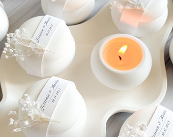 May include: White, spherical candles with a ribbon and floral accent, arranged on a white tray. One candle is lit, showing a warm orange glow. Each candle has a tag that reads "Jessenia & Martin 10.11.2025".