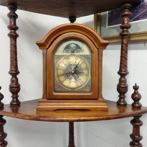 May include: A wooden antique clock with a decorative face and a brown wooden frame. The clock is displayed on a corner shelf with ornate, turned supports. The shelf is made of dark wood and has a curved edge.