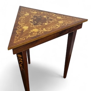 May include: A triangular wooden side table with a dark brown finish. The tabletop features an inlaid floral design in lighter wood tones. The table has three slender legs, each with decorative accents, and is suitable for a living room or entryway.