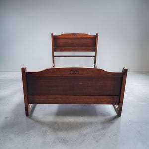 May include: A dark brown wooden bed frame featuring a tall headboard and a lower footboard. Both the headboard and footboard have a curved top edge with a small, centred decorative carved detail. The frame is constructed with solid wood panels.