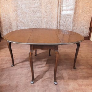 May include: Oval wooden dining table with a dark brown finish. The table has four curved legs and a silver trim around the edge. The table is approximately 152 cm long and 102 cm wide.
