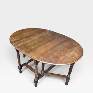 May include: An oval drop-leaf table crafted from medium-toned brown wood with a visible grain pattern. The table features a smooth tabletop surface and is supported by four turned legs connected by a sturdy wooden stretcher base frame.