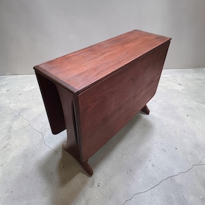 May include: A rectangular drop-leaf table crafted from dark brown wood with a smooth, polished surface. The table features two hinged side panels that hang vertically, supported by a central base structure with curved legs on each end.