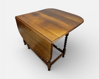 May include: A wooden drop-leaf table with a warm brown finish. The table has a rounded rectangular top and a single leaf that is folded down. The legs are dark brown and have a turned design. The table is set against a white background.