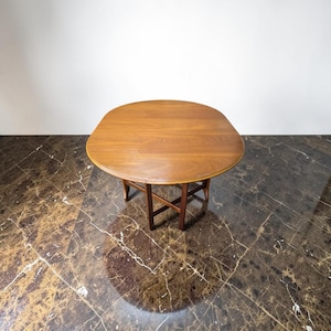 May include: A round wooden table with a dark brown finish. The table has a rounded top and four legs. The table is set against a white wall and a brown marble floor. The table is approximately 76 cm in diameter.