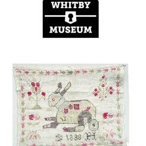 Op de afbeelding: Een witte linnen sampler met een kruissteekontwerp van een konijn, twee bomen en de datum "1888".